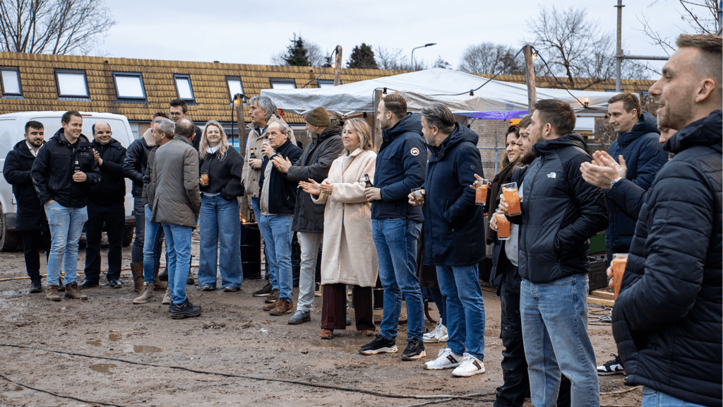 Ondernemers en betrokkenen staan samen buiten op de bouwplaats van een bedrijfspand in Arnhem tijdens het bouwfeest aan de Overmaat. De groep applaudisseert en houdt drankjes vast terwijl zij luisteren naar een spreker, met op de achtergrond de bouwlocatie en tijdelijke overkappingen.