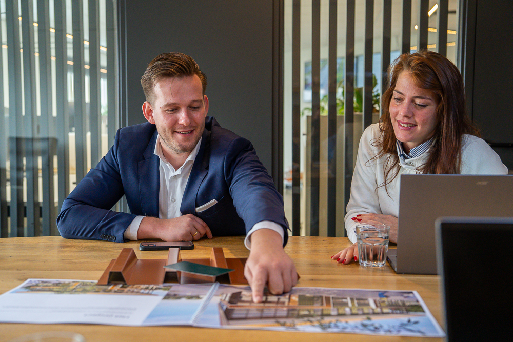 Twee collega’s bespreken tijdens een overleg het project in Ochten (het huis dat binnenkort te koop komt), zittend aan een vergadertafel met een brochure en projectvisualisaties voor zich. Eén collega wijst details aan op de plannen, terwijl de ander meekijkt op een laptop. De setting straalt aandacht, samenwerking en zorgvuldige voorbereiding van dit woonproject in Ochten uit.