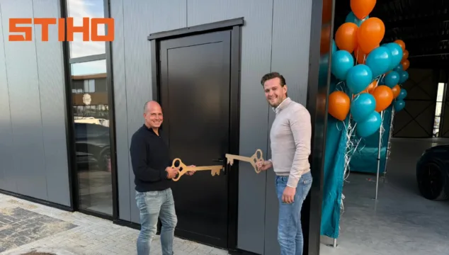 Twee mannen openen feestelijk de deur van het nieuwe Stiho-pand in Nijmegen met grote houten sleutels, naast een rij oranje en turquoise ballonnen. De gevel van het moderne gebouw is zichtbaar op de achtergrond. Zoekterm: Stiho Nijmegen.