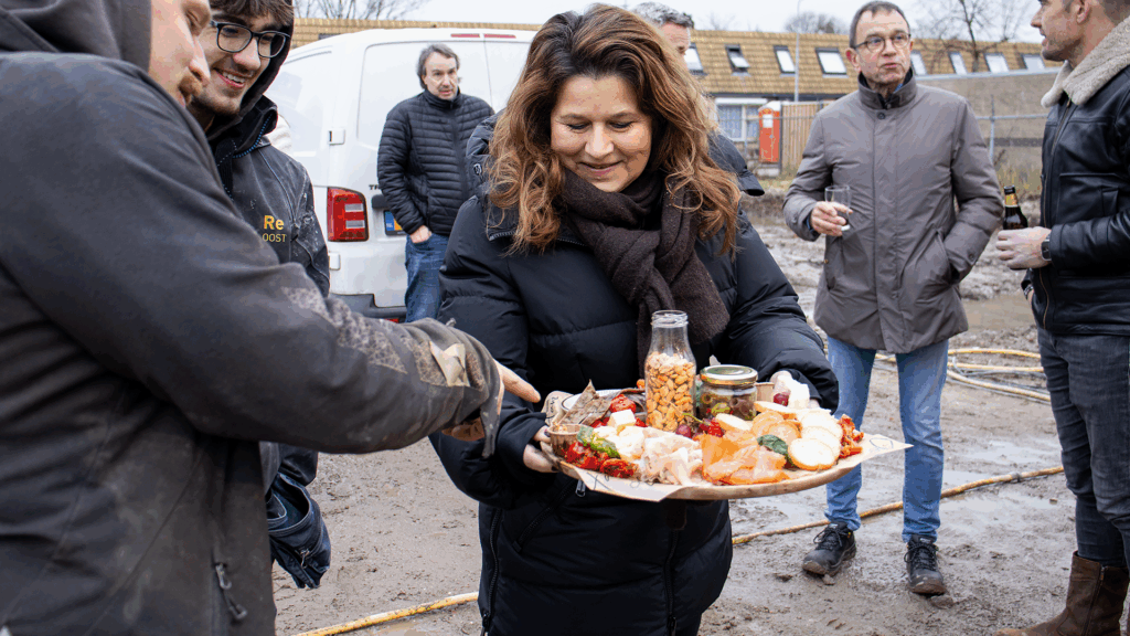 Ondernemers en betrokkenen genieten van een borrelplank tijdens het bouwfeest van het bedrijfspand in Arnhem aan de Overmaat, staand op de bouwplaats met modderige ondergrond en in gesprek over de voortgang van het project.