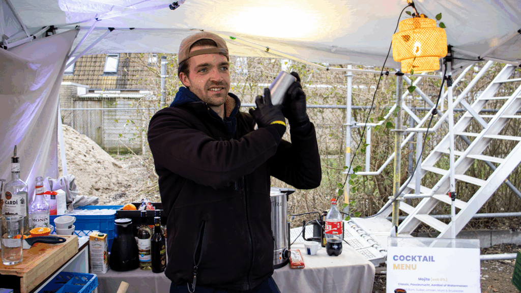 Barman die cocktails mixt onder een partytent tijdens het bouwfeest van het bedrijfspand in Arnhem aan de Overmaat, met een cocktailmenu op de bar en de bouwplaats zichtbaar op de achtergrond.