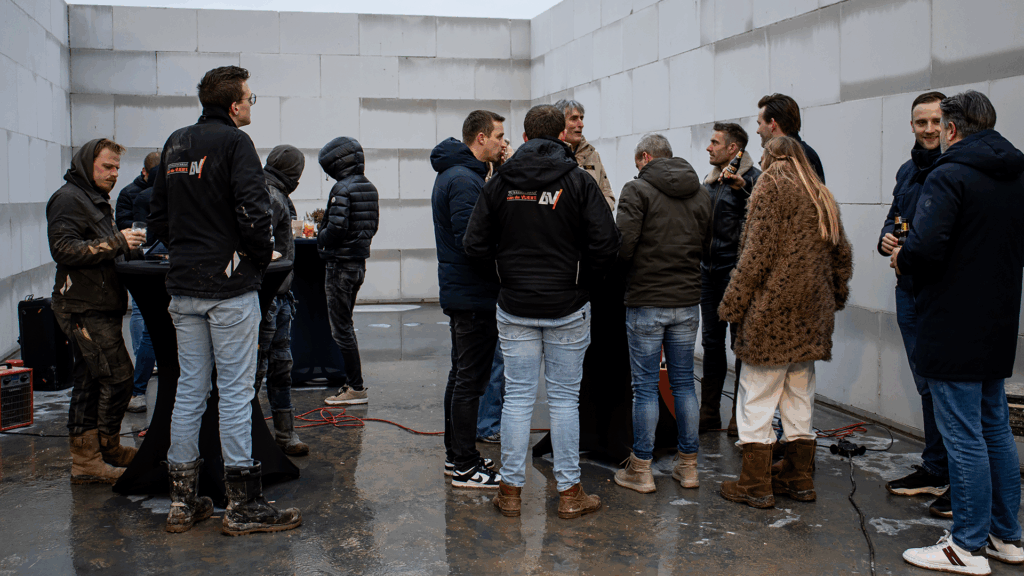 Ondernemers en bouwpartners in gesprek tijdens het bouwfeest van het bedrijfspand in Arnhem aan de Overmaat, staand in het nog niet wind en waterdichte casco pand rond statafels met drankjes, terwijl de binnenmuren in aanbouw zichtbaar zijn.