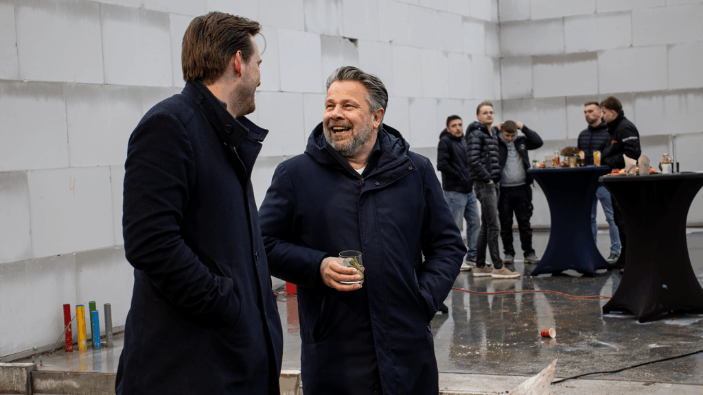 Twee ondernemers in gesprek tijdens het bouwfeest van het bedrijfspand in Arnhem aan de Overmaat, staand op de bouwplaats met een drankje in de hand, terwijl op de achtergrond andere gasten bij statafels samenkomen in het nog niet wind en waterdichte pand.