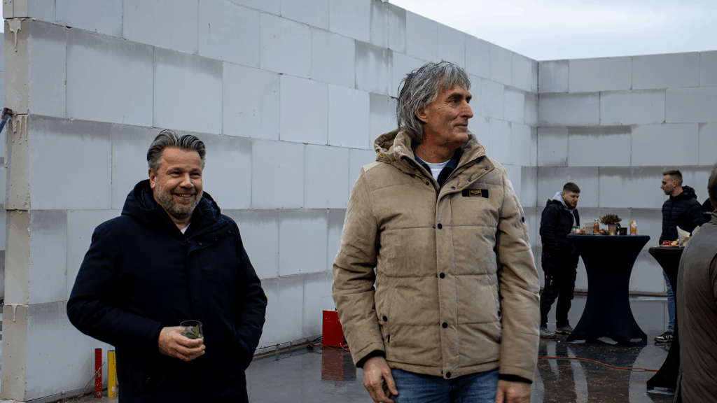Twee ondernemers lopen lachend rond tijdens het bouwfeest van het bedrijfspand in Arnhem aan de Overmaat, terwijl op de achtergrond gasten bij statafels staan in het nog in aanbouw zijnde casco pand met zichtbare kalkzandsteenmuren.