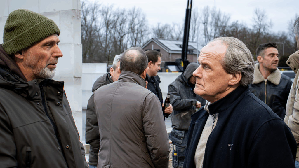 Ondernemers in gesprek tijdens het bouwfeest van het bedrijfspand in Arnhem aan de Overmaat, staand op de bouwplaats tussen de opgetrokken kalkzandsteenmuren terwijl op de achtergrond andere gasten met drankjes samenkomen.