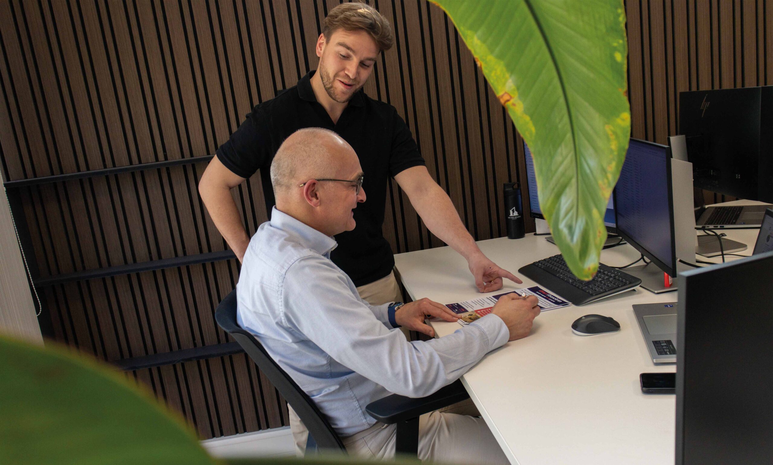 Twee collega’s bespreken aan een bureau de investeringsmogelijkheden voor een bedrijfspand in Ammerzoden, met computerschermen, een brochure en groene plant op de voorgrond.