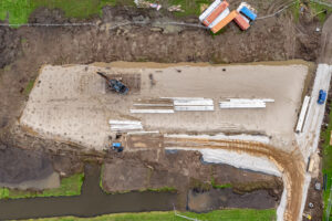 Luchtfoto van de bouwplaats van het nieuwbouw bedrijfspand in Bodegraven aan de Tolnasingel, met zicht op heipalen, bouwmaterialen en funderingswerkzaamheden in volle gang.