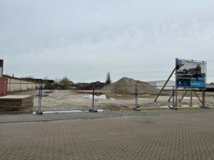 Leeg terrein met zandhopen en bouwbord achter hekwerk: Bouwlocatie voor fase 1 van bedrijfspand Oudenbosch, voorbereidingen gestart op strategische locatie dichtbij Breda.