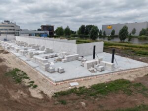 Bouwplaats van een bedrijfspand in Bodegraven met gemetselde wanden in opbouw, stapels bouwmaterialen en zicht op omliggende bedrijven en waterpartij.